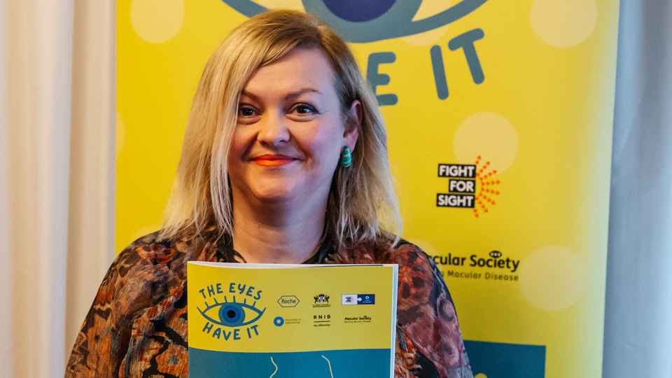 Woman with blonde hair holding up The Eyes Have It poster at Westminster Eye Health Day.