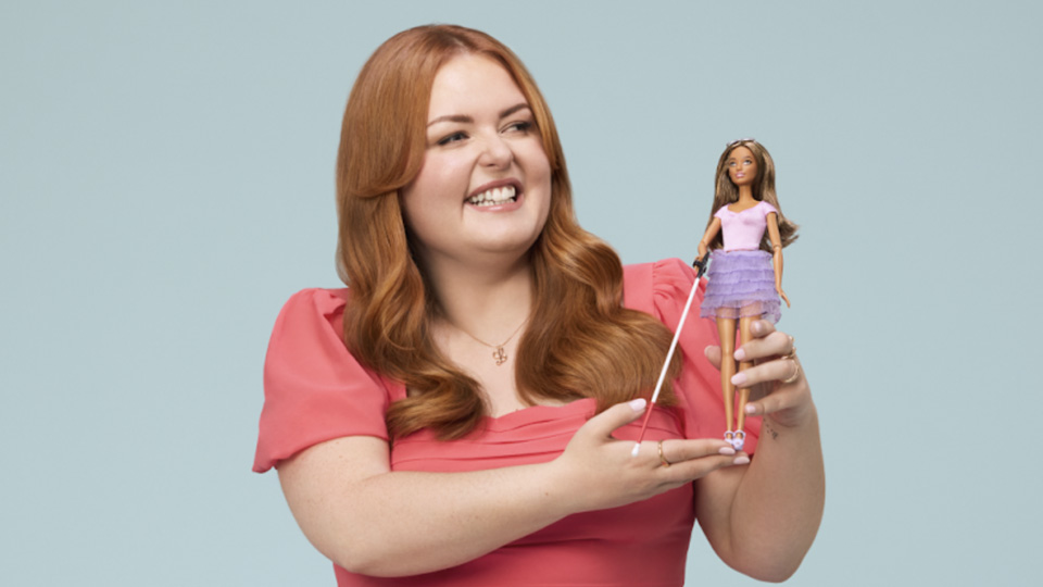 A woman with red hair and a salmon-coloured dress holds a barbie doll in her hands. The barbie doll has long brown hair, a white cane, a purple tutu style skirt and pink t-shirt.