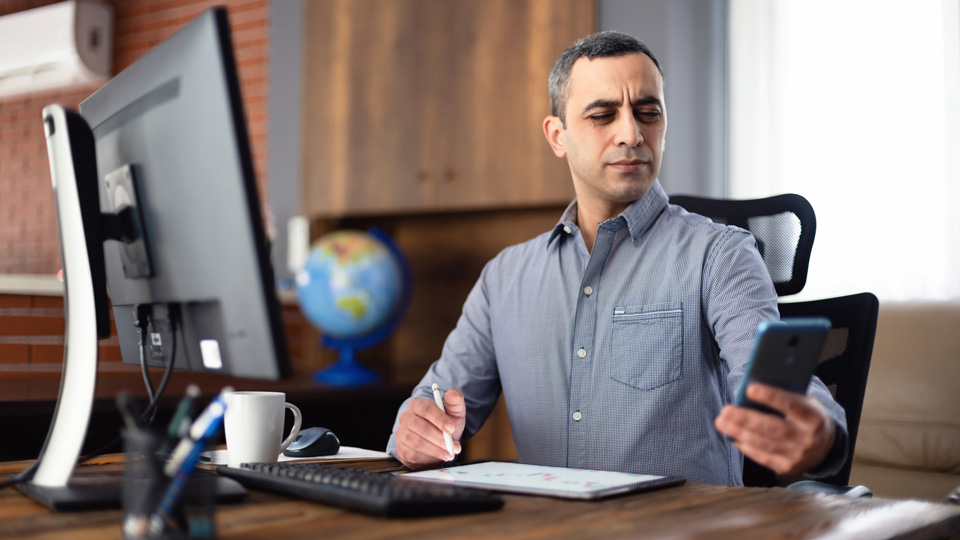 A man in his early 40s sits at a desk in front of a large computer screen and squints as he tries to read the screen of his smartphone 