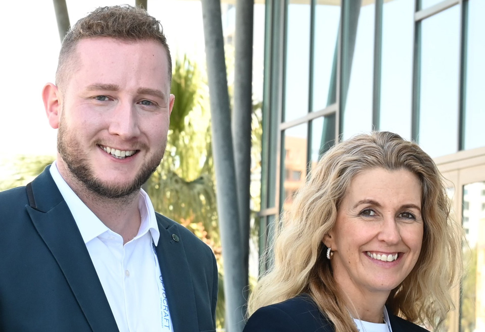 Tom (left) and Sarahjane (right) stand together, smiling in the sunshine, both wearing smart blazer jackets. In the background is the glass front of an office building