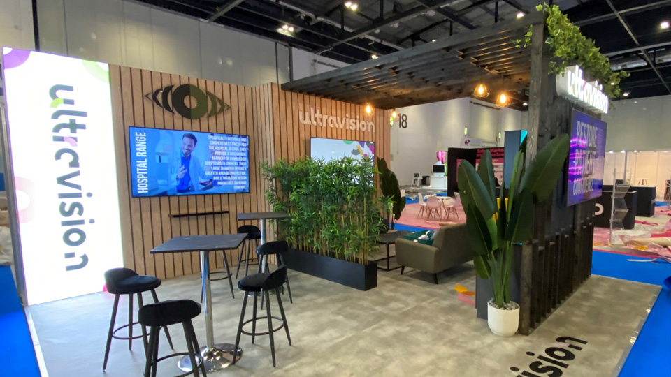 Ultravision’s stand at 100% Optical combines dark wood panelling with plants. The new logo – a geometric eye and bold black text with green and purple elements, is presented on a bright light panel 