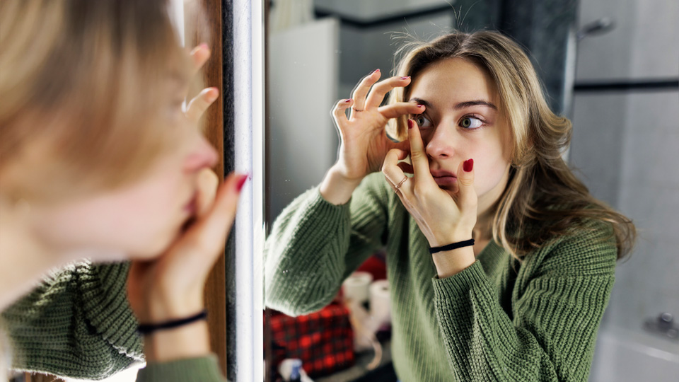 A girl in a green jumper is applying a contact lens in front of a bathroom mirror  