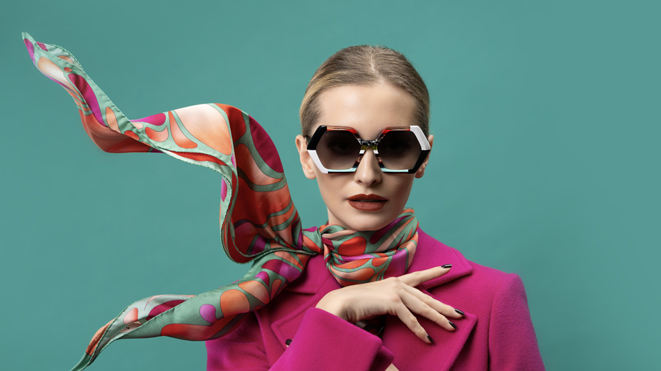 A woman wears a bright pink coat with a silk scarf in a green, peach, red and pink pattern. She wears oversized hexagonal sunglasses with strips of light blue, orange, black and white