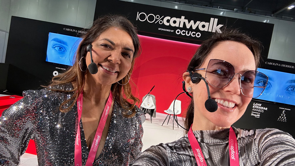 Dr Rachna Murthy and Amy Gallant Sullivan pose wearing microphones on the 100% Catwalk. Rachna wears a silver glittery dress, while Amy wears a black and white dress of pop art style eyes 