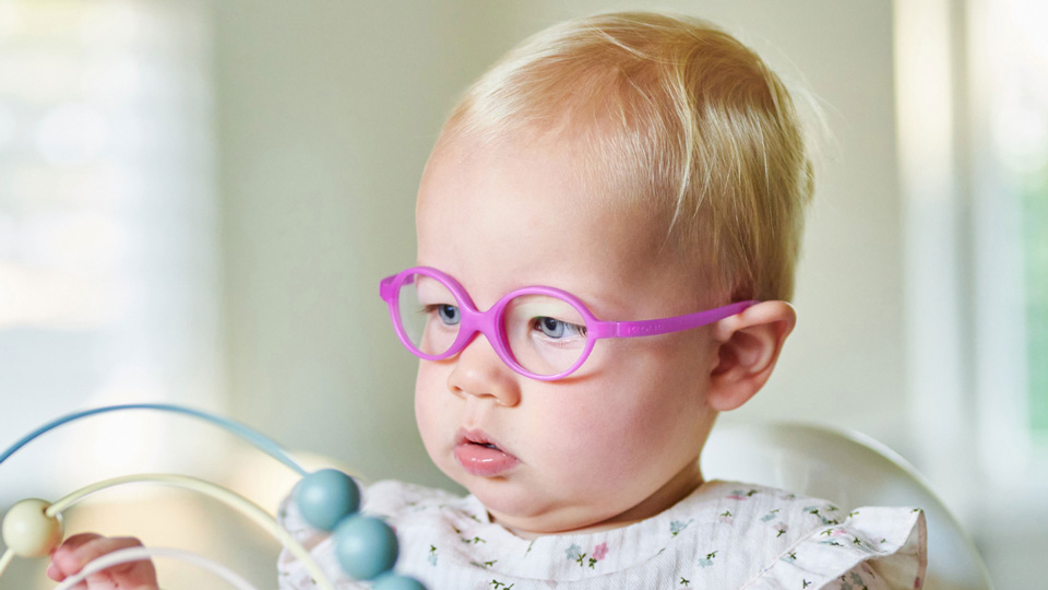 An infant girl sits in a high chair focusing intently on a toy, she wears bright pink glasses