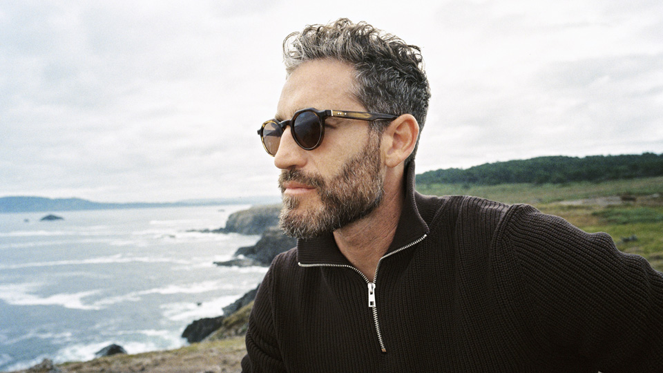 A man wearing brown pantos sunglasses looks off-frame at the ocean. The coastline curves away behind him