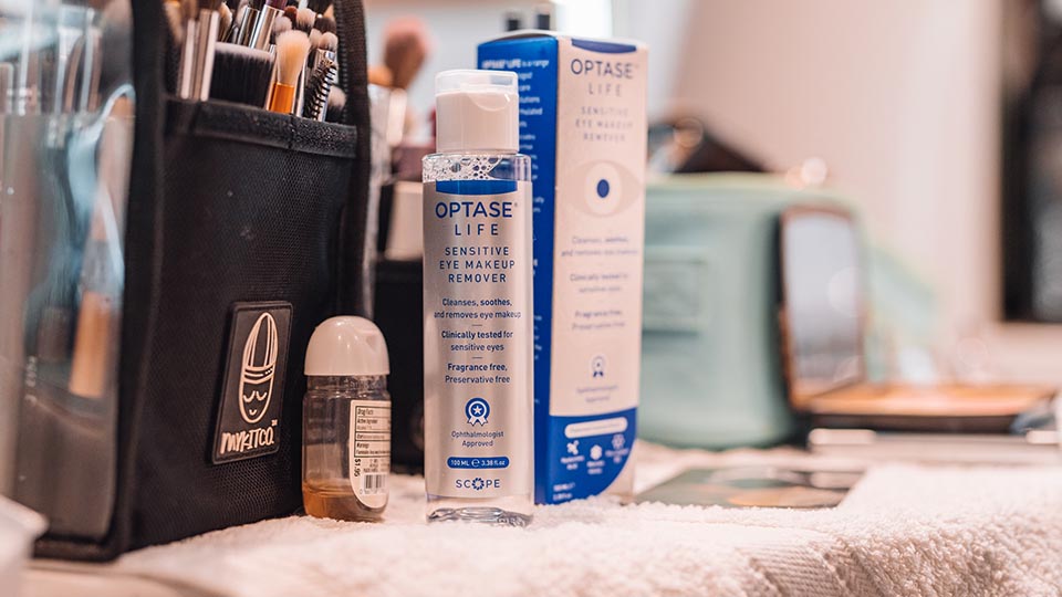 A bottle of Optase Life stands on a dresser next to its box. Both have silver and blue branding and the liquid in the bottle is clear. In the foreground is a large black makeup bag full of makeup brushes of varying sizes. Another makeup bag and palette are blurred in the background