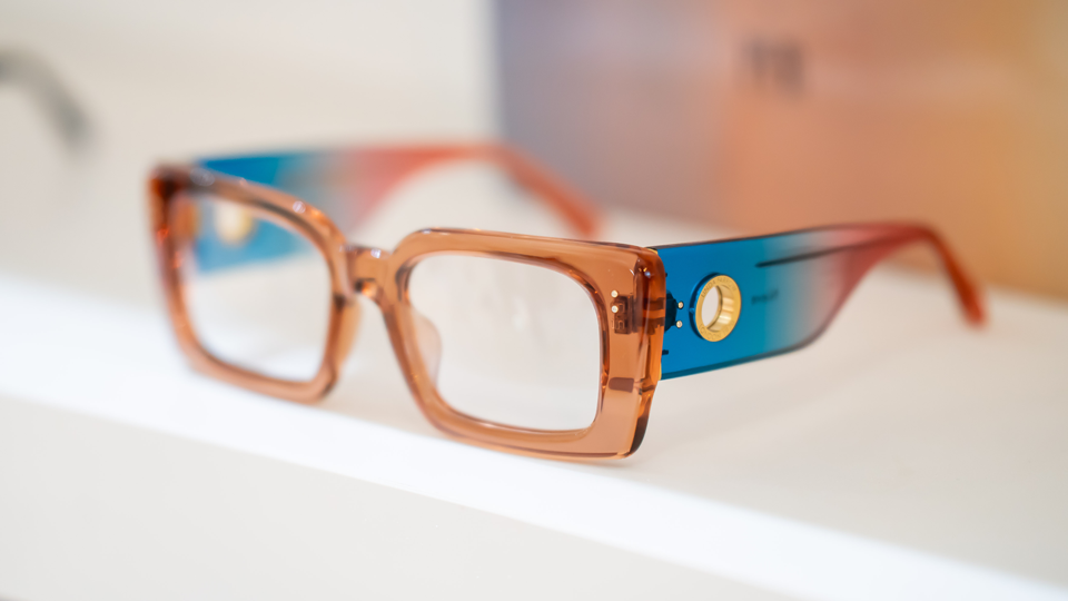 A pair of chunky acetate frames rest on a white shelf. The frames are transluscent, with a peach front, and sides that transition from blue to a deep pink