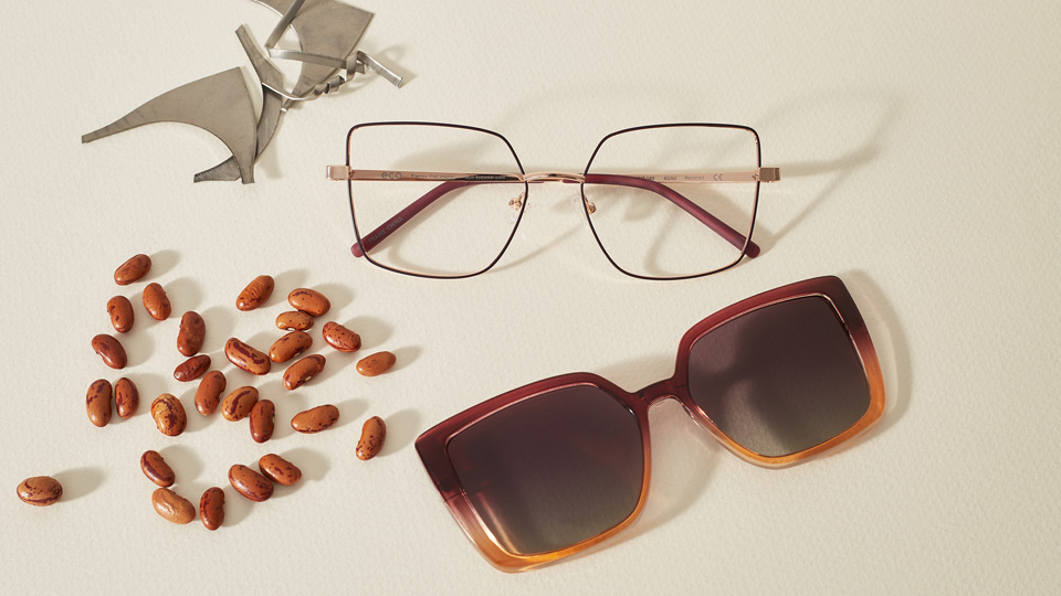 Two frames laid next to a scattered handful of beans and a pile of scrap recycled metal. The squared cat’s eye frame is made of fine metal with pink end tips, and the clip-on sunglasses front ombres from purple-pink to brown-orange