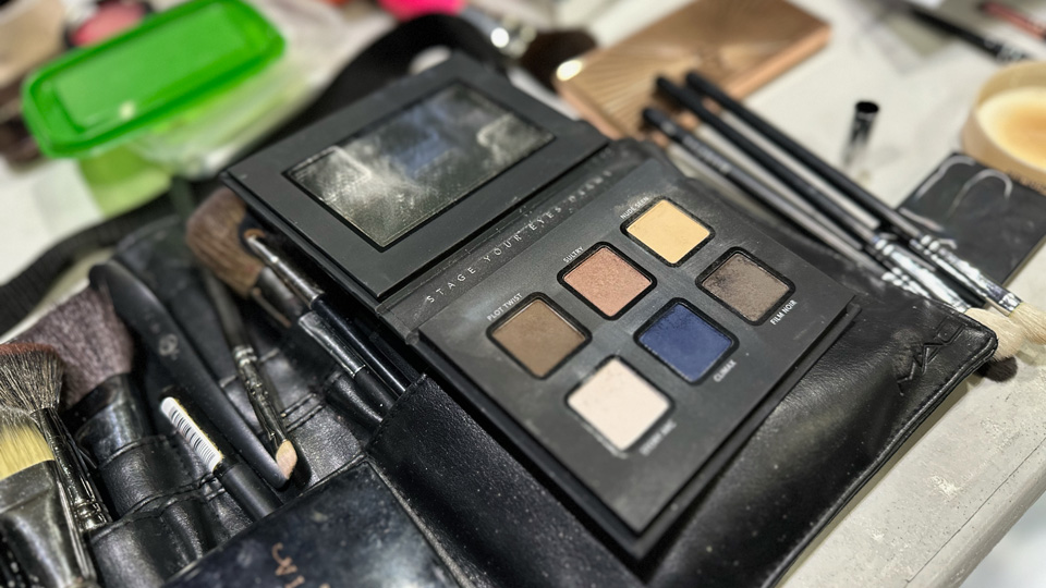Make-up is laid out across a table, clearly mid-use. The Eyes Are The Story eyeshadow palette is open with shades displayed ranging from a brown titled ‘Plot Twist’ to a black-grey called ‘Film Noir’