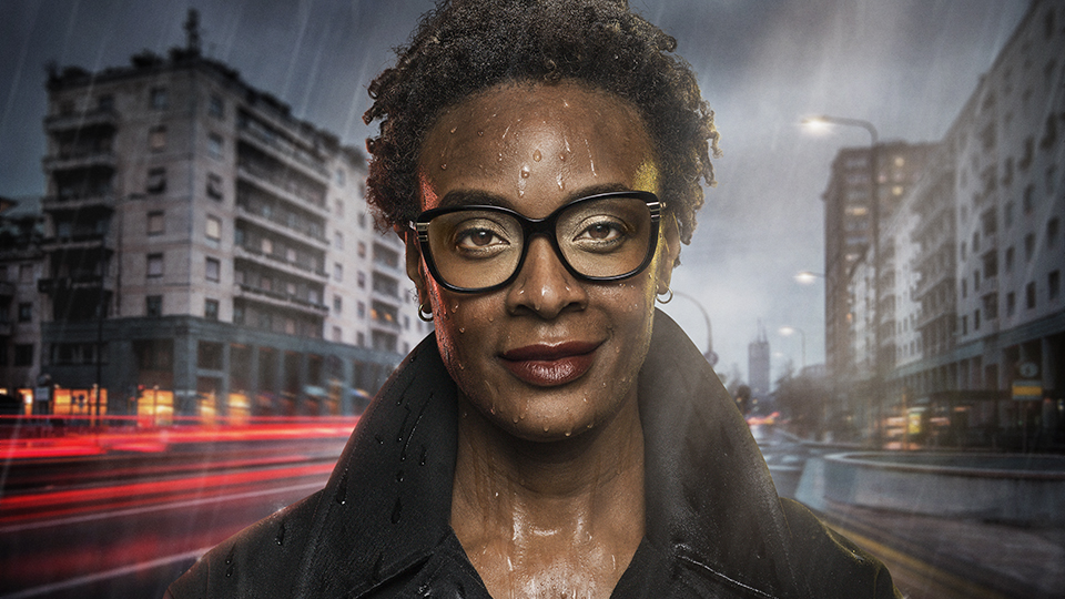 A black woman wearing glasses stands in the rain at night, with a blurred city backdrop  