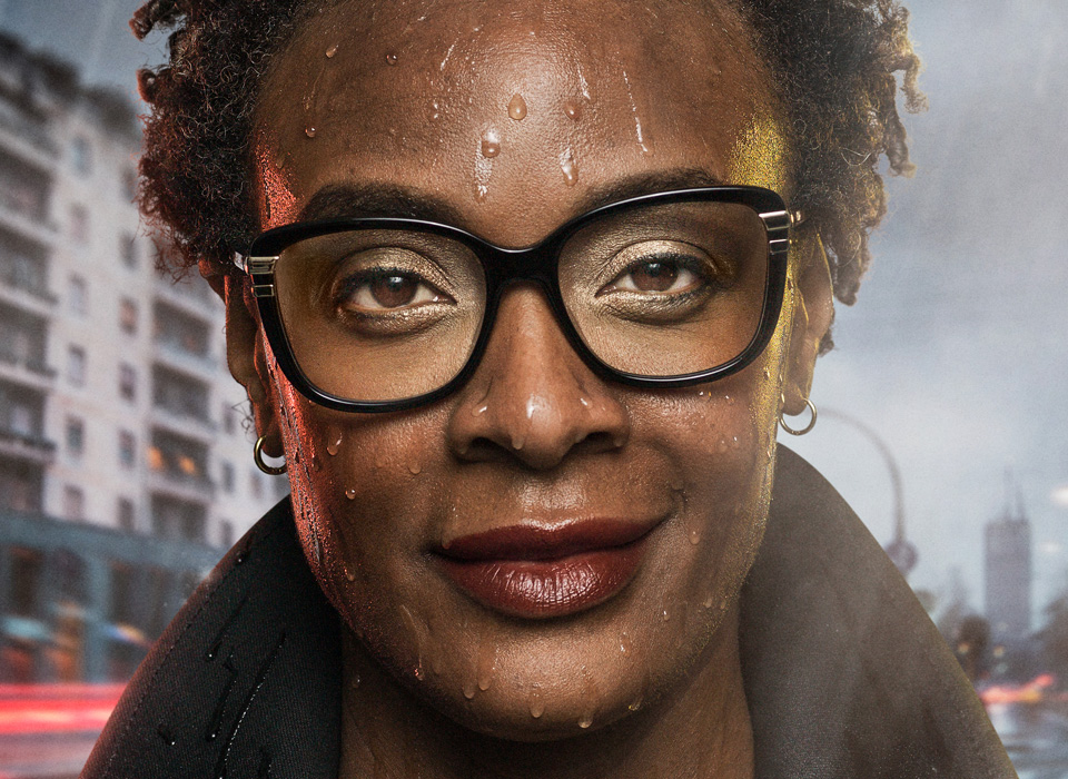 A woman faces the camera, wearing glasses and smiling despite the rain pouring down around her. In the background is a scene of a city at night in the rain