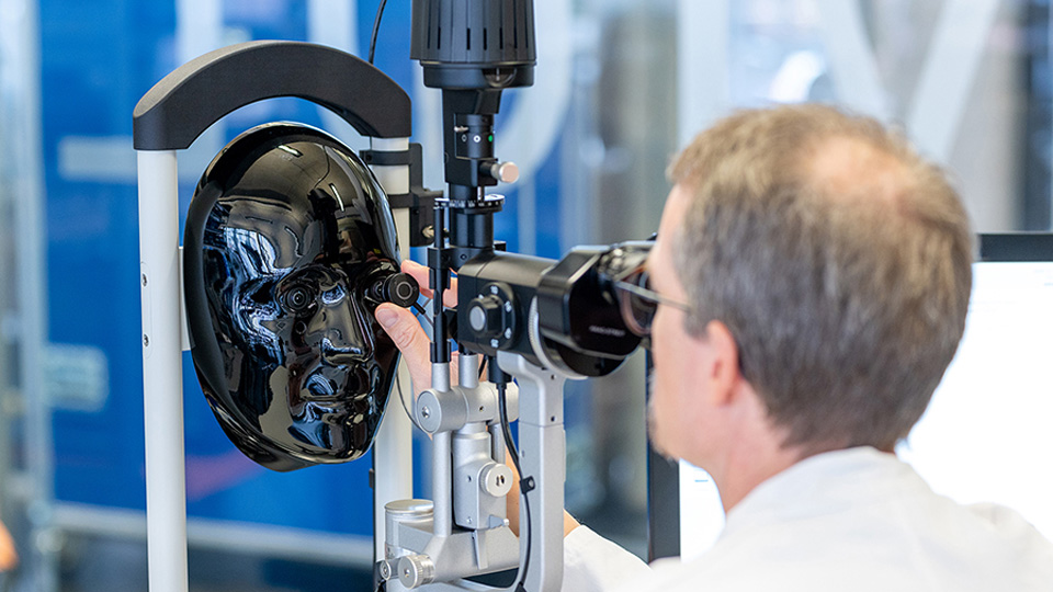 A man in a white lab coat has his eye up to the slit lamp device, holding an instrument up to a shiny plastic model face