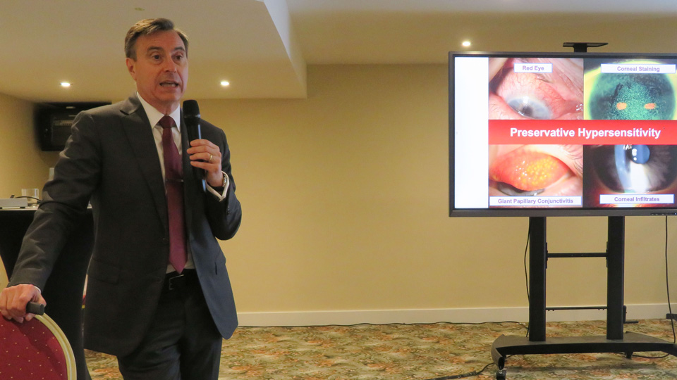 A man in a dark suit and mauve tie speaks into a microphone while leaning on a chair. Behind him is a large screen displaying the words ‘preservative hypersensitivity’ and a series of pictures of everted eyelids
