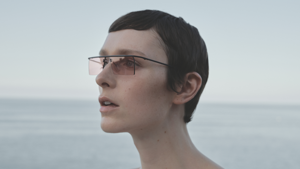 A woman with a pixie cut stands against the backdrop of the ocean wearing small rectangle rimless frames with soft pink lenses