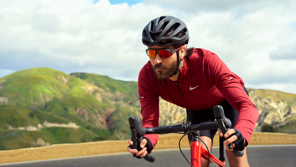  A cyclist wears red and leans close to the handlebars. He wears sporty sunglasses with red lenses and a black helmet. Green hills roll away behind him