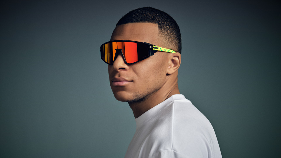 Kylian Mbappé poses in a pair of Oakley glasses with yellow temples and orange lenses