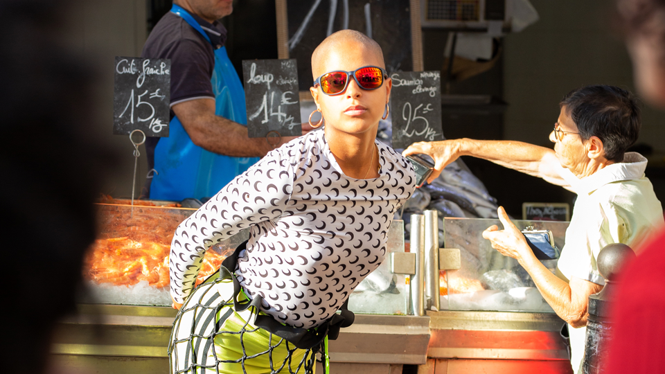 A woman poses wearing dark sunglasses, and a skirt made of a wide square net, behind her is a fish stall