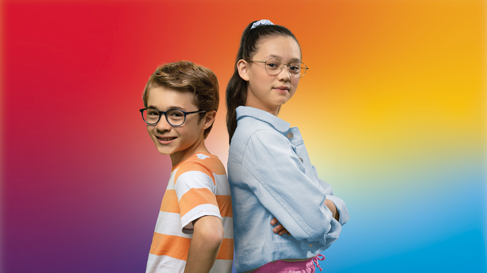 Two children, a boy and girl, stand with their arms crossed and confident looks wearing spectacles. They stand in front of a rainbow background 