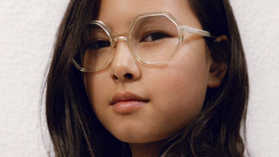  A young girl looks towards the camera, wearing clear octagonal-shaped frames 