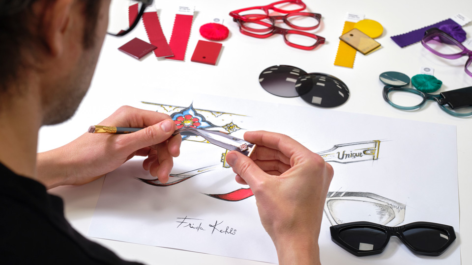 A photograph taken over the shoulder of an eyewear designer, who is holding the front of a pair of frames in their hands over a sketch, with material swatches and other designs laid out 