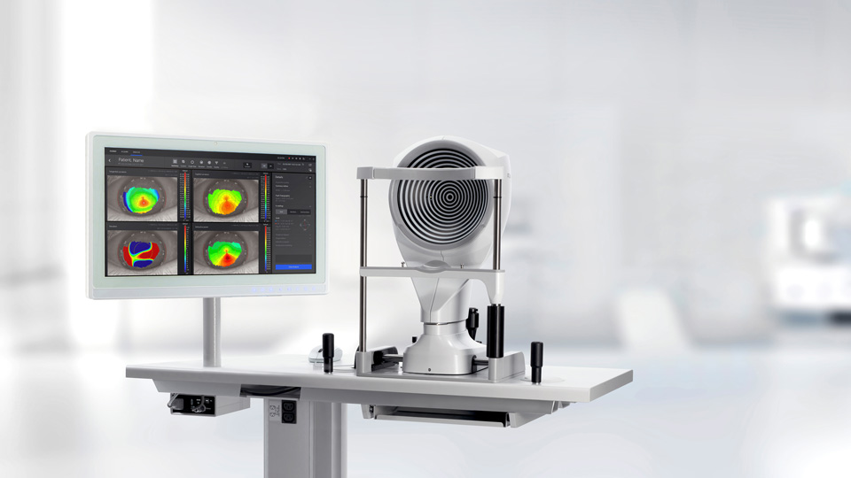 A device is positioned towards the camera, looking into the concentric circles of the instrument. The screen displays heat-style maps of an eye. The background is blurred but is white and seems clinical
