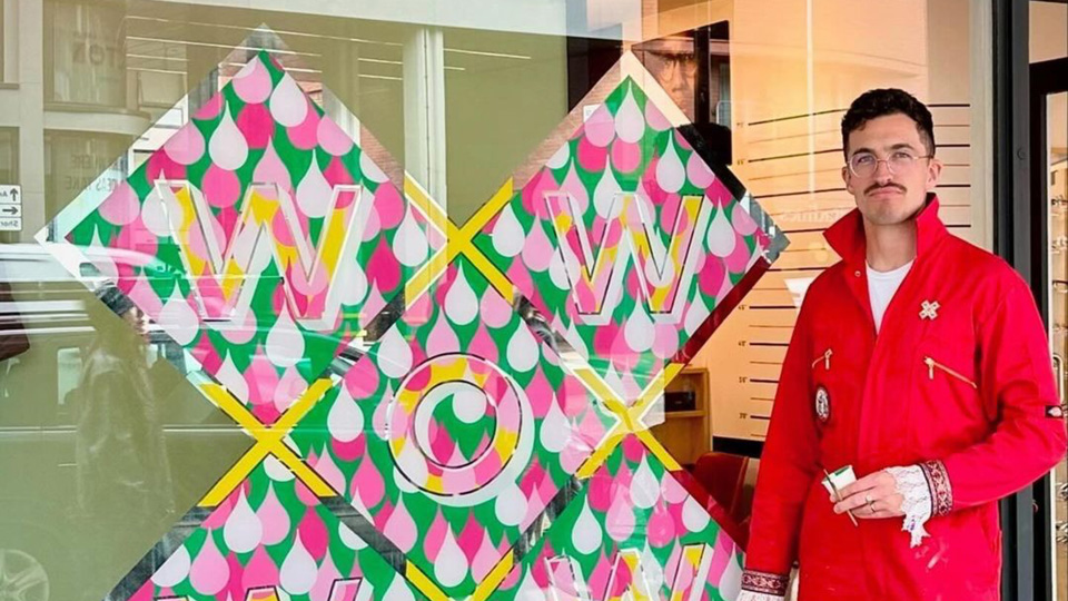Archie, wearing bright red overalls that have cuffs trimmed with lace, stands beside the window of Cult Vision, which has a colourful mural spelling the word ‘WOW’ across an X shape. Motifs of pink raindrops are used to symbolise spring. 
