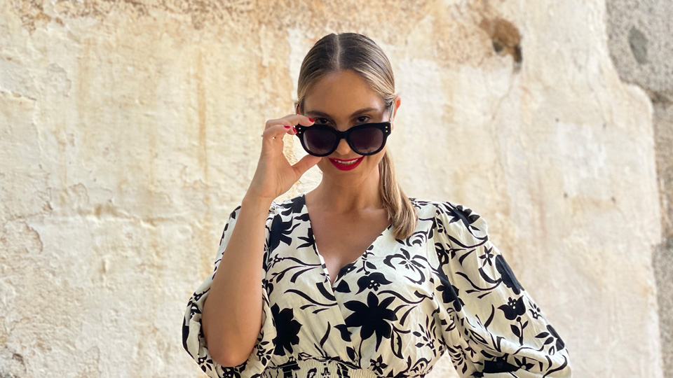 Carolina is outside in the sunshine wearing a black and white floral summer dress, she is peeking over the top of her square black sunglasses 