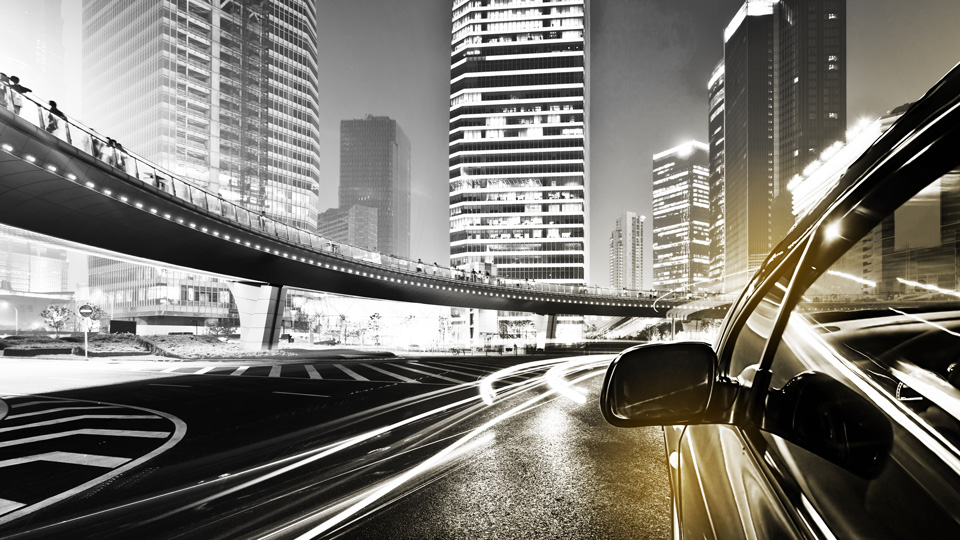 A wing mirror of a car is visible driving down a busy road. The black and white image is a long exposure so the headlights of other cars streak across the image like light trails 