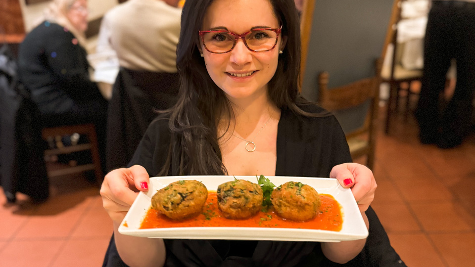 Clare Brent wears bright red frames in a rectangle shape. She is in a restaurant and holds up a plate of food 