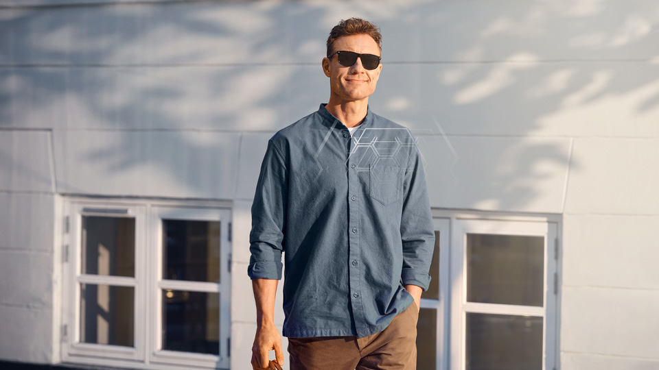A man stands outside in casual clothes and sunglasses, a pale outline of lens technology is overlaid on the image, indicating the technology in the lenses