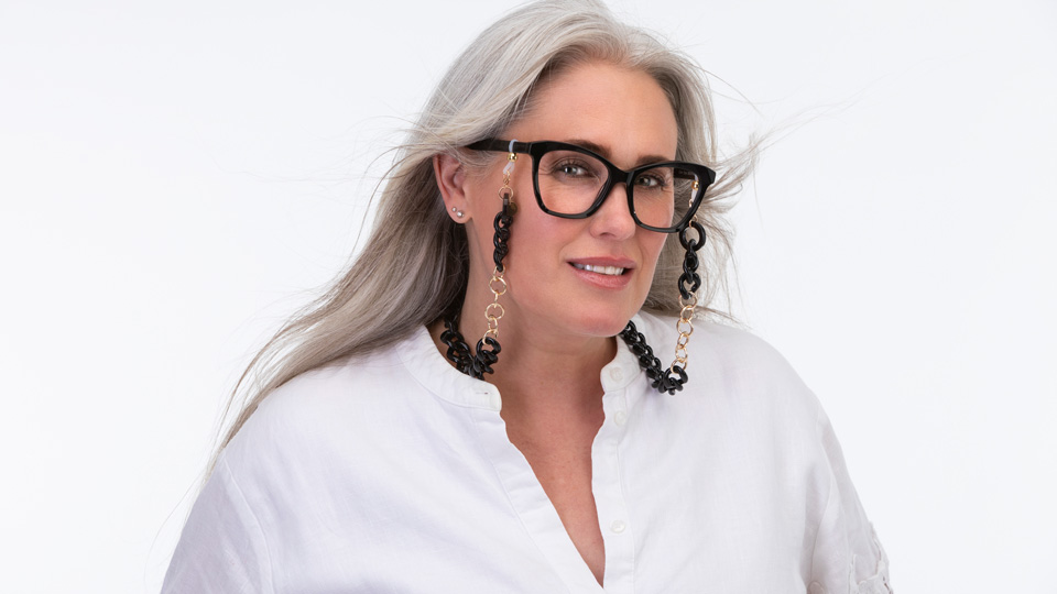 A woman wearing white stands against a white backdrop. She wears chunky black spectacles with a bold glasses chain made up of black recycled acrylic links and sleek aluminium chains