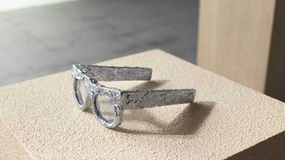 A frame rests on a mottled concrete plinth. The frame is a silver colour and very chunky, with straight arms, the texture appears rough and ready, inspired by the tin foil model this frame was based on