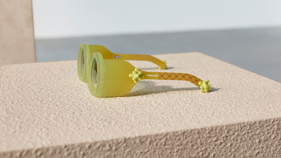 A frame rests side-on to the camera. The frame is a soft, semi-transluscent yellow in a gradient from almost light-green to a darker yellow. The arm of the frame is studded with two plastic flower elements, ‘Yinka Ilori’ is eteched in white against the arm, which has a simplistic red flower pattern