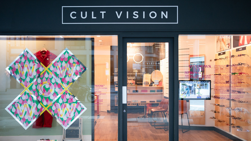 The window of Cult Vision displays Archie Proudfoot’s ‘WOW’ design, an extravaganza of light and dark pink raindrops on a green background, with the letters picked out in fine lines of yellow and shining silver. Directly behind the painting, Archie has hung his iconic red painting overalls, and left his stepladder and used paintpots. In the background, the practice is lit with a warm yellow light.
