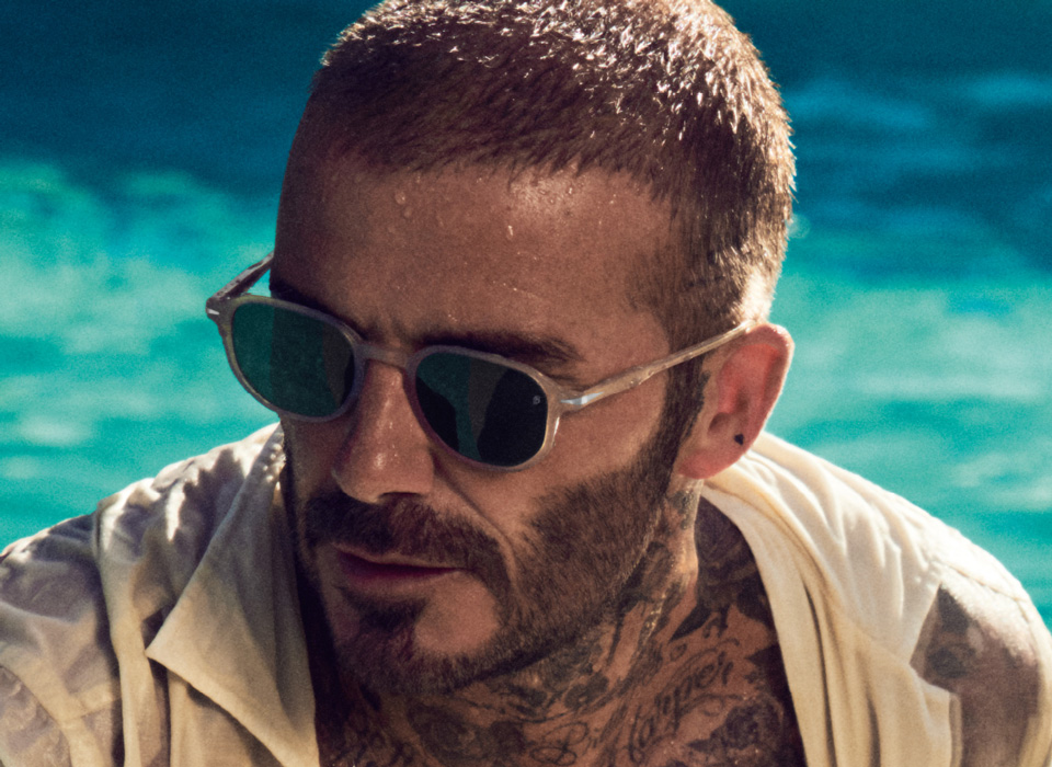 David Beckham wears a pair of sandy colours sunglasses from his collection in a summery pool-side image 