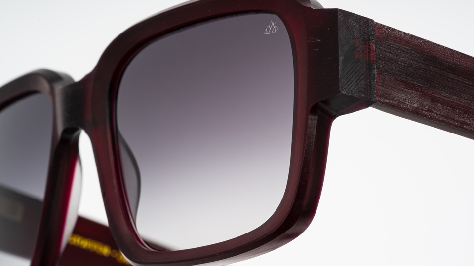 A close up of one corner of a chunky red pair of sunglasses. The top corner of one of the dark lenses has a small etching of a geometric mountain 