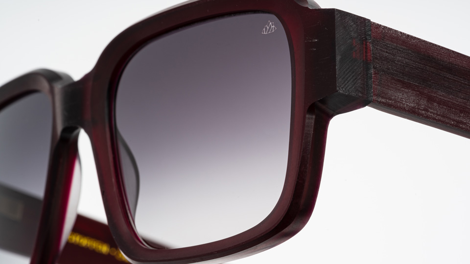 A close up of one corner of a chunky red pair of sunglasses. The top corner of one of the dark lenses has a small etching of a geometric mountain