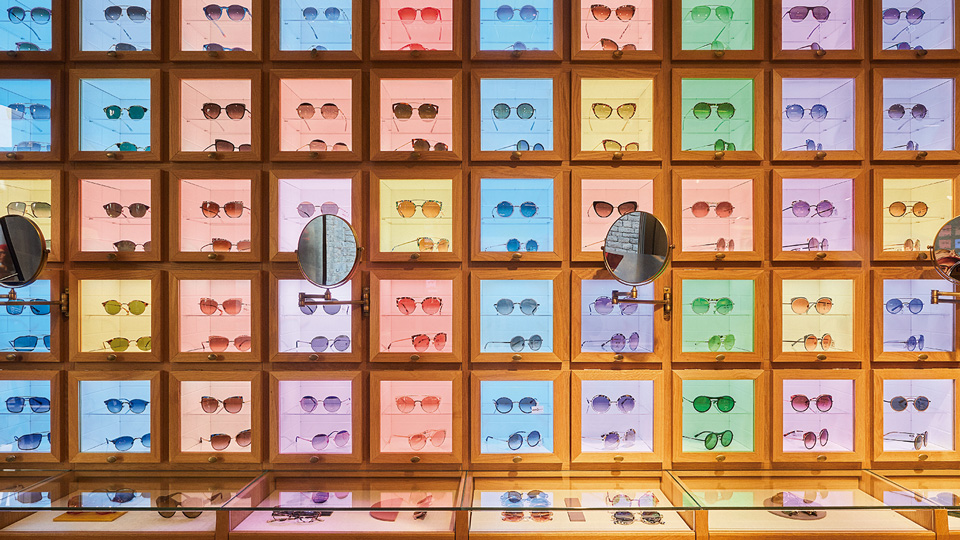 A wall divided into cubes, each with a coloured light of eight pink, yellow, purple, blue or green. Each cube has two pairs of sunglasses. Three mirrors are placed on moveable arms halfway up the wall. Display cabinets line the base of the walls 