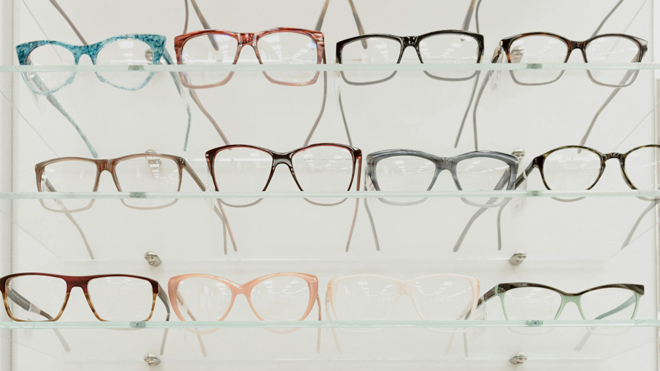 Rows of spectacles are arranged on a glass display cabinet with a mirror in the background