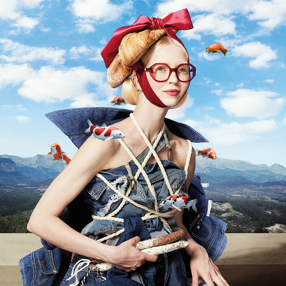 A young blonde woman is posed like a model in a renaissance-era painting. She wears large red frames and a large red bow of a matching colour. Croissants are photoshopped as her hair. She appears to wear a collage of denim wrapped in fishing rope, as goldfish swim in open air around her. The background is a mountain range with white clouds on a blue sky