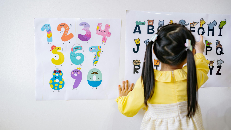 A little girl has her back to the camera and is running her finger over a wall poster with the alphabet, different animals are associated with each letter