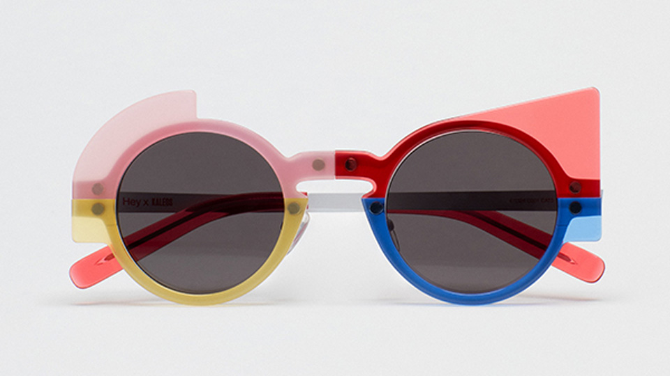 A woman with dark hair in a pixie cut wears round sunglasses with four modular sections – a red cat’s eye, a rounded blue base, a squared yellow, and round white base.  