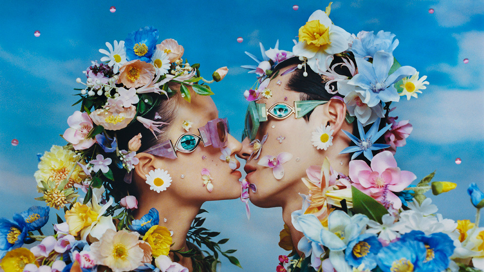 A young man and woman lean in to kiss, they both wear chunky acetate frames that feature a giant eye on the sides. She wears pink frames and his are a teal green. Both appear to be overgrown with an array of flowers including daffodils, roses, giant daisies and lilies. The image has a soft, vintage-style quality 