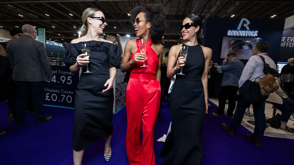 Three women, two in black dresses and one in a red jumpsuit, walk towards the camera holding glasses and laughing with each other. They each wear sunglasses: two wear bold black frames with detailing and the third pair is a butterfly shape with metal sides 