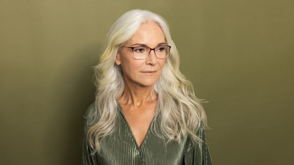  A woman with white hair wears a shiny sage green dress and stands against an olive green background. She wears spectacles with a dark top and sides that transitions into a clear plastic at the bottom of the frame 