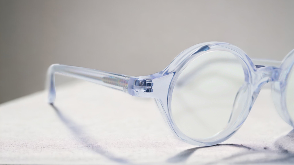 A clear acetate frame is angled so only the left lens and arm are displayed. The side of the frame is slightly broader, to provide coverage for a thicker lens.  