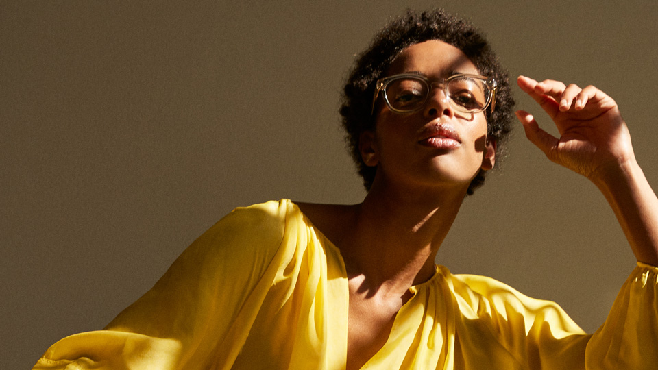 A woman wears clear square spectacles and wears a sunshine yellow outfit, the folds and drapes of the material catching the light and shade 