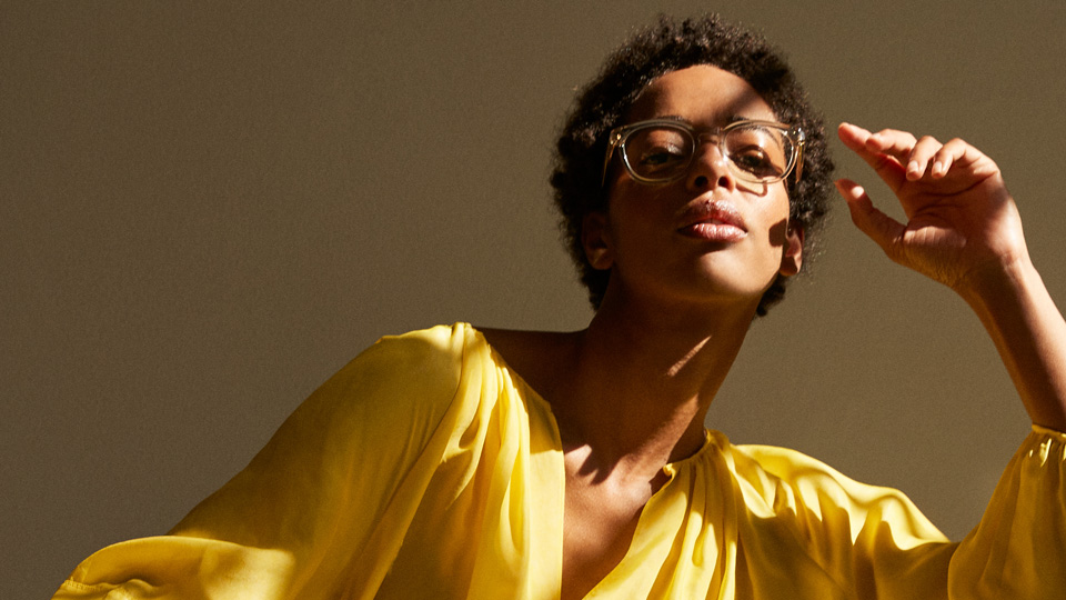 A woman wears clear square spectacles and wears a sunshine yellow outfit, the folds and drapes of the material catching the light and shade