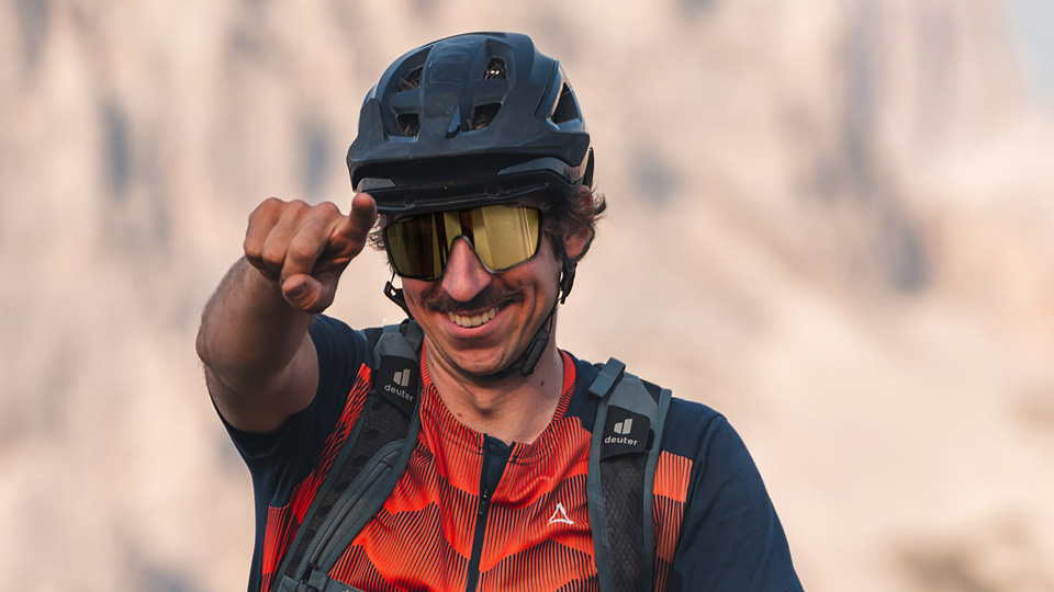 Gerald smiles, holding up the peace pose, on a bike in a mountainous area of rolling peaks and lush greenery 